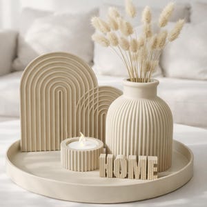 Könnte beinhalten: Ein beigefarbenes Dekotablett mit einer Vase mit Trockenblumen, zwei bogenförmigen Skulpturen, einem Kerzenhalter mit einer brennenden Kerze und dem Wort "HOME". Die Objekte sind in einer neutralen Farbpalette gehalten und schaffen eine minimalistische Ästhetik.