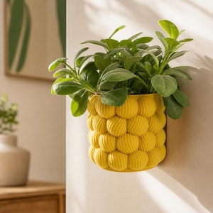 Op de afbeelding: Een felgele wandplantenbak met een getextureerd, bubbelachtig oppervlak met herhalende ronde bollen. De plantenbak bevat een weelderige groene plant met brede, gladde bladeren die naar boven en naar buiten uit de opening steken.