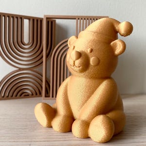 Könnte beinhalten: Eine goldfarbene, 3D-gedruckte Bärenfigur mit Schlafmütze, die vor zwei rechteckigen Rahmen mit konzentrischen Bogenmustern steht. Der Bär hat eine glatte, abgerundete Form mit sichtbaren Schichtlinien aus dem Druckprozess.