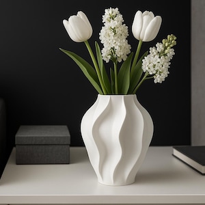Könnte beinhalten: Eine weiße Keramikvase mit wellenförmigem Design, gefüllt mit weißen Tulpen und Hyazinthen. Die Vase steht auf einem weißen Tisch, daneben eine dunkelgraue Box und ein schwarzes Buch. Der Hintergrund ist eine dunkelgraue Wand.