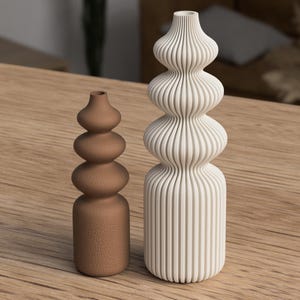 Könnte beinhalten: Zwei dekorative Vasen. Eine ist eine hohe, cremefarbene Vase mit vertikalen Rillen und einer bauchigen Form. Die andere ist eine kleinere, braune Vase mit einem ähnlichen, aber glatteren, bauchigen Design. Beide Vasen stehen auf einer Holzoberfläche.
