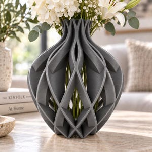 Könnte beinhalten: Eine dunkelgraue Vase mit einer komplexen, offenen geometrischen Gitterstruktur und sich wiederholenden rautenförmigen Ausschnitten. Die Vase hat einen schmalen Hals, der sich zu einem runden Körper weitet und weiße Blumen sowie grünes Blattwerk enthält.