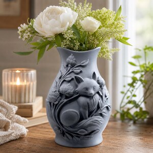 Könnte beinhalten: Eine matte hellgraue Keramikvase mit einem erhabenen Reliefdesign eines Fuchses, umgeben von Blattwerk und Blütenknospen. Die Vase ist mit einem Strauß aus weißen Pfingstrosen und kleinen grünen Füllblumen gefüllt, was ein strukturiertes Blumenarrangement ergibt.