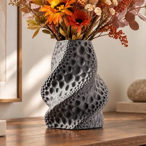 Può includere: Un vaso grigio scuro con una silhouette a spirale ritorta e una superficie strutturata caratterizzata da numerose rientranze circolari. Il vaso è riempito con una vivace composizione di fiori secchi nei toni dell'arancione, giallo e rosso.