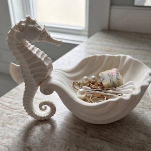Könnte beinhalten: Eine mattweiße Keramikschale in Form einer Muschel mit einer aufrechten Seepferdchenfigur an der Seite. Die Schale enthält verschiedene kleine Gegenstände, darunter goldfarbene Ringe, perlenartige Ohrstecker und weiße Wattestäbchen.