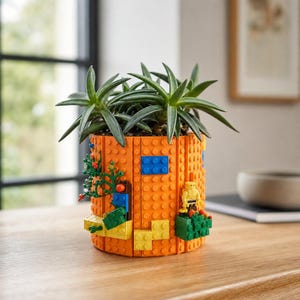 Könnte beinhalten: Ein zylindrischer Pflanztopf aus orangefarbenen Kunststoff-Bausteinen. Die Außenseite ist mit kleinen dekorativen Elementen versehen, darunter eine gelbe Figur, grüne Pflanzenteile und blaue rechteckige Akzente. Eine grüne Sukkulente befindet sich im Inneren.