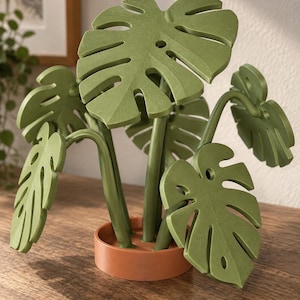 Könnte beinhalten: Eine dekorative Pflanzenskulptur mit mehreren grünen Monstera-Blättern, die charakteristische Ausschnitte und Perforationen aufweisen. Die Blätter sind an grünen Stielen befestigt, die in einer runden, terrakottafarbenen Basis verankert sind.