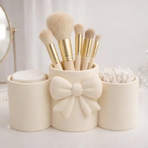 Könnte beinhalten: Ein cremefarbener Kosmetik-Organizer aus Keramik mit drei zylindrischen Fächern. Das mittlere Fach ist mit einer erhabenen Schleife verziert. Er enthält fünf Make-up-Pinsel mit goldenen Zwingen, weiße Wattepads und weiße Wattestäbchen.