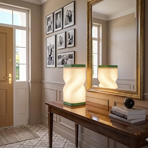 Könnte beinhalten: Eine moderne, beleuchtete Lampe mit einem einzigartigen, gedrehten Design. Die Lampe hat einen weißen, ziehharmonikaartigen Schirm mit grüner Umrandung. Sie steht auf einem dunklen Holztisch, neben einem Stapel Bücher und einer dekorativen Kugel. Der Raum hat einen Spiegel und gerahmte Fotos.