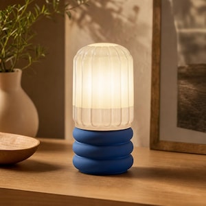 Può includere: Una lampada da tavolo con un paralume cilindrico in vetro bianco smerigliato a coste verticali che emette una luce calda. La base è composta da tre anelli in ceramica blu opaco impilati e arrotondati, creando un netto contrasto materico.