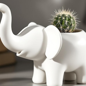 Könnte beinhalten: Ein weißer, elefantenförmiger Übertopf aus Keramik mit glatter, matter Oberfläche. Der Elefant ist mit erhobenem Rüssel dargestellt, und ein kleiner, runder grüner Kaktus mit spitzen Dornen ist in die Öffnung auf seinem Rücken gepflanzt.