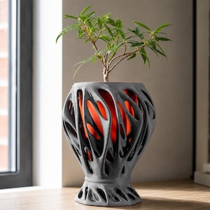 Peut inclure: Un cache-pot gris de forme organique avec un extérieur complexe en forme de toile. Le cache-pot présente une doublure intérieure orange vif visible à travers les découpes de la coque extérieure grise. Une petite plante verte est à l'intérieur.