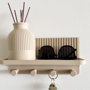 Könnte beinhalten: Ein beiges Wandregal mit gerippter Rückwand und vier zylindrischen Haken. Eine gerippte beige Vase mit dunkelbraunen Schilfstäbchen steht neben einer schwarzen Sonnenbrille auf dem Regal. Ein Metallschlüsselring hängt an einem der Haken.
