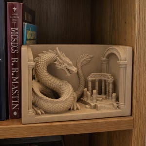 Könnte beinhalten: Ein beigefarbener Buchständer mit einem skulptierten Drachen, der sich neben einer Miniatur-Ruine mit Säulen und einem Torbogen windet. Der rechteckige Sockel trägt das detaillierte Relief, das auf einem Holzregal neben Büchern steht.