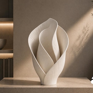 Op de afbeelding: Een sculpturale gebroken witte vaas met een gelaagd, organisch ontwerp dat lijkt op een bloeiende bloem. Het oppervlak van de vaas vertoont fijne, verticale ribbeltexturen over de gehele gebogen, overlappende, bloembladachtige vormen.