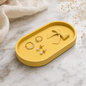 Peut inclure: Un plateau ovale jaune avec un détail de nœud en relief sur la surface. Le plateau contient une bague dorée avec une petite perle, une bague dorée avec un anneau de perles, et une paire de boucles d'oreilles dorées avec des perles blanches.