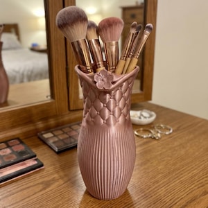 Könnte beinhalten: Ein roségoldener Make-up-Pinselhalter mit gesteppter Textur im oberen Bereich und vertikalen Rillen im unteren Teil. Der Halter ist am Rand mit einer kleinen Blumenverzierung versehen und enthält mehrere Make-up-Pinsel.