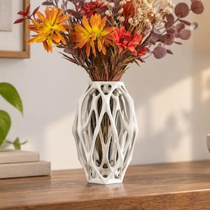 Könnte beinhalten: Eine weiße Vase mit einer komplexen, offenen gitterartigen geometrischen Struktur. Die Vase hat einen sechseckigen Boden und ein spitz zulaufendes, käfigartiges Design, das einen klaren Glaseinsatz mit einem Strauß getrockneter Blumen in Orange, Rot und Braun zeigt.