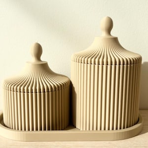 Op de afbeelding: Twee beige cilindrische houders met verticale geribbelde texturen en bijpassende deksels, elk bekroond met een kleine bolvormige knop. De houders hebben verschillende hoogtes en staan samen op een bijpassend ovaalvormig dienblad.
