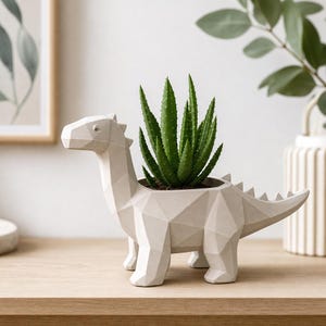 Könnte beinhalten: Ein hellbeiger, geometrischer Dinosaurier-Pflanztopf mit facettiertem, kantigem Design. Der hohle Rücken des Dinosauriers enthält eine leuchtend grüne Aloe-Pflanze mit spitzen, gezackten Blättern. Der Topf steht auf vier stabilen Beinen.