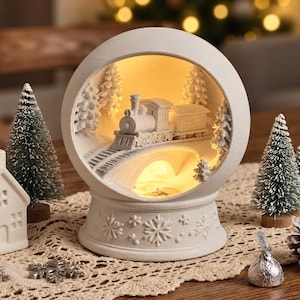 Peut inclure: Une lampe décorative blanche circulaire présentant une scène intérieure avec un train traversant un pont au milieu d'une forêt. La base est ornée de motifs de flocons de neige en relief et l'intérieur est éclairé par une lueur douce.