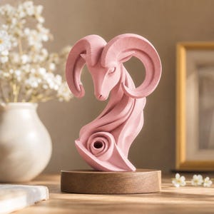 Puede incluir: Una escultura de color rosa mate de una cabeza de carnero con grandes cuernos curvados, con un cuello estilizado que se transforma en un patrón de remolino circular en la base. La escultura está montada sobre una base circular de madera.