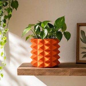 Könnte beinhalten: Ein leuchtend orangefarbener zylindrischer Übertopf mit einer geometrischen, pyramidenförmigen Texturoberfläche. Der Topf enthält eine üppige grüne Efeutute mit panaschierten herzförmigen Blättern. Das Gefäß zeigt ein Muster aus Spitzen.
