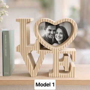 Könnte beinhalten: Eine beige, gerippte, dreidimensionale Wortskulptur, die das Wort LOVE bildet, wobei der Buchstabe O durch einen herzförmigen Bilderrahmen ersetzt ist, der ein Schwarz-Weiß-Porträt eines lächelnden Paares enthält. Das Objekt ist als Model 1 beschriftet.