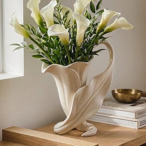 Könnte beinhalten: Eine cremefarbene Keramikvase in Form einer Calla-Lilie mit einer weit ausgestellten Öffnung und einem geschwungenen, blattartigen Fuß. Die Vase ist mit einem Strauß weißer Calla-Lilien und grünem Blattwerk gefüllt, was ihr eine skulpturale Optik verleiht.