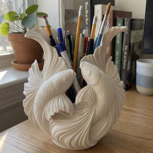 Pode incluir: Um organizador de secretária cor de creme em forma de dois peixes koi com barbatanas fluidas. A escultura apresenta escamas detalhadas e barbatanas caudais curvas que formam um recipiente central. Está cheio de várias canetas e lápis.