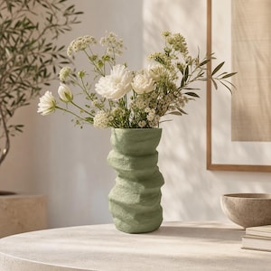 Könnte beinhalten: Eine salbeigrüne Keramikvase mit einer texturierten, unebenen und organisch geschichteten Oberfläche. Die Vase enthält ein großes Arrangement aus weißen Blumen, darunter Pfingstrosen und zarte Füllblüten, mit dünnen Stielen und grünen Blättern.