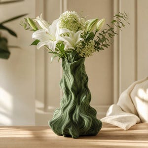 Könnte beinhalten: Eine dunkelgrüne Keramikvase mit einer texturierten, wellenförmigen und organischen Oberflächenstruktur. Die Vase ist mit einem Strauß aus weißen Lilien, kleinen hellgrünen Blütenclustern und dünnen grünen Eukalyptuszweigen gefüllt.