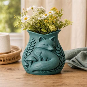 Könnte beinhalten: Eine blaugrüne Vase mit einem Relief eines Fuchses, der sich zwischen Farnen und Pilzen zusammenrollt. Die Vase ist mit einem Strauß aus weißen Gänseblümchen und kleinen gelben Blumen gefüllt und bildet ein Dekorationsobjekt mit matter Oberfläche.