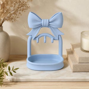 Può includere: Un supporto portagioie azzurro caratterizzato da un grande fiocco decorativo sulla parte superiore, un arco curvo con tre piccoli ganci per appendere oggetti e una base a vassoio ovale liscia per riporre piccoli accessori.