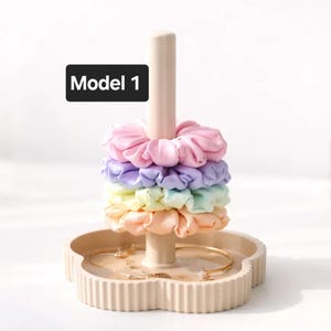 May include: A beige jewelry stand with a flower-shaped base and a central post holding four pastel-colored fabric scrunchies in pink, purple, yellow, and peach. A black rectangular label with the white text Model 1 is visible above the scrunchies.