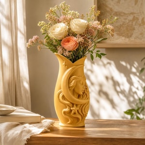 Puede incluir: Un jarrón de cerámica color amarillo mostaza con un relieve de un rostro enmarcado por una luna creciente y estrellas. El jarrón contiene un ramo de rosas blancas y color melocotón mezcladas con pequeñas flores rosas y follaje verde.