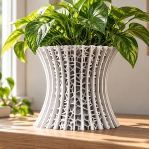 Könnte beinhalten: Ein weißer 3D-gedruckter Übertopf mit einer ausgestellten, sanduhrförmigen Silhouette und einem komplexen, geometrischen Gittermuster auf der Außenseite. Der Übertopf enthält eine leuchtend grüne Efeutute mit panaschierten Blättern.