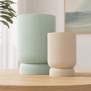 Könnte beinhalten: Zwei zylindrische Pflanzgefäße mit vertikaler Rippenstruktur. Das größere Gefäß ist hell mintgrün, das kleinere ist neutral beige. Beide verfügen über einen abgerundeten Sockelbereich, der etwas breiter als der Hauptkörper ist.