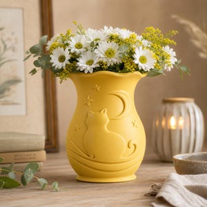 Könnte beinhalten: Eine gelbe Vase mit gewelltem Rand und einem Relief, das eine Katze unter einer Mondsichel und Sternen zeigt. Die Vase ist mit einem Strauß aus weißen Margeriten, gelben Goldrutenblüten und grünen Eukalyptusblättern gefüllt.
