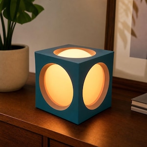 Könnte beinhalten: Eine würfelförmige Lampe in Blaugrün mit kreisförmigen Aussparungen an den Seiten und auf der Oberseite. Jede Öffnung gibt den Blick auf eine warm leuchtende, kugelförmige Lichtquelle frei, die die geometrische Form des Objekts betont.