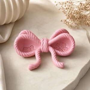May include: A light pink decorative tray shaped like a bow with a twisted rope texture. The center of each bow loop features a small, embossed floral pattern. The object has a smooth, glossy finish and a symmetrical design with two hanging ends.