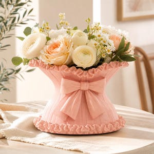 Könnte beinhalten: Eine hellrosa Keramikvase mit einem ausgestellten, gerüschten Rand und Boden sowie einer markanten, skulpturalen Schleife auf der Vorderseite. Die Vase ist mit einem Strauß aus weißen und pfirsichfarbenen Ranunkeln gefüllt.