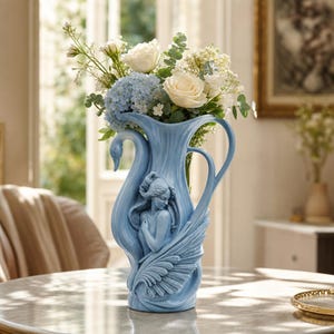 Könnte beinhalten: Eine hellblaue Keramikvase mit einem skulpturalen Relief einer geflügelten Figur und einem Henkel in Schwanenhalsform. Die Vase ist mit einem Blumenstrauß aus weißen Rosen, blauen Hortensien und grünem Blattwerk gefüllt.