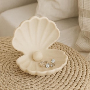 Könnte beinhalten: Eine cremefarbene, geöffnete muschelförmige Schmuckschale mit einem glatten, kugelförmigen, perlenartigen Akzent in der Mitte. Zwei silberfarbene Ringe mit klaren, facettierten Steinen liegen in der unteren Hälfte der Muschelschale.