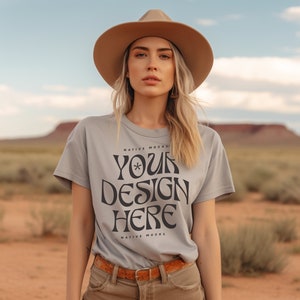May include: A woman wearing a light gray t-shirt with the text "YOUR DESIGN HERE" printed on the front. The shirt is a mockup for a design project. The woman is wearing a brown hat and is standing in a desert landscape.