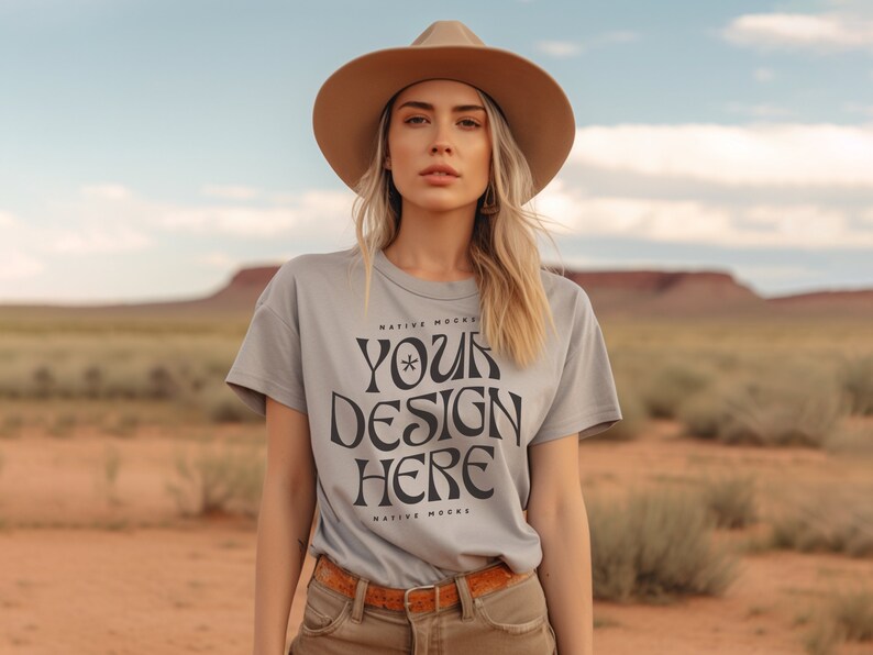 May include: A woman wearing a light gray t-shirt with the text "YOUR DESIGN HERE" printed on the front. The shirt is a mockup for a design project. The woman is wearing a brown hat and is standing in a desert landscape.