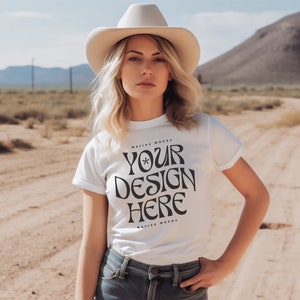 May include: A woman wearing a white t-shirt with the text "YOUR DESIGN HERE" printed on it. She is wearing a cowboy hat and standing in a desert landscape.
