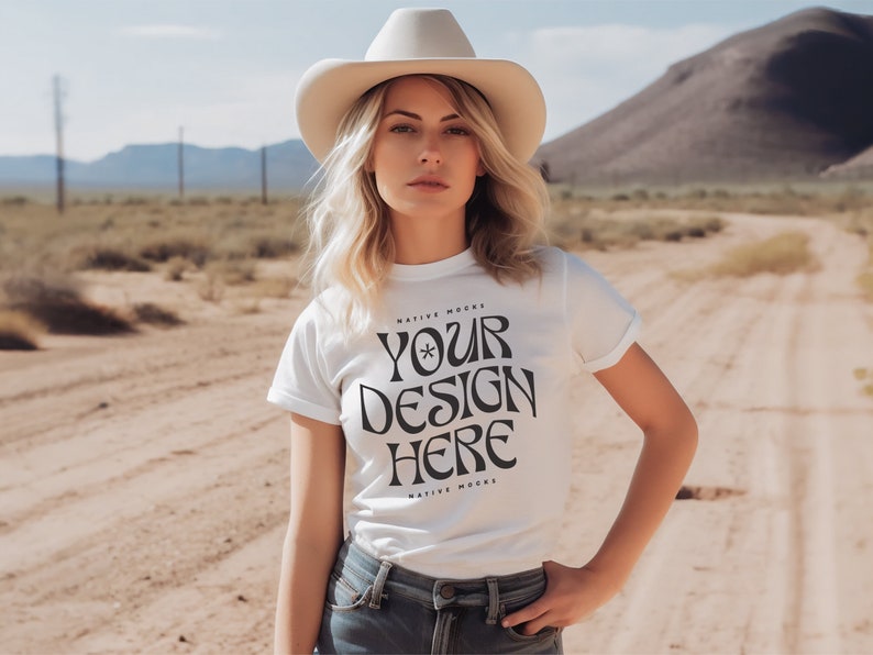 May include: A woman wearing a white t-shirt with the text "YOUR DESIGN HERE" printed on it. She is wearing a cowboy hat and standing in a desert landscape.