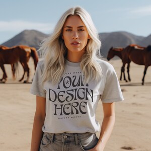 May include: A woman wearing a light gray t-shirt with the text "YOUR DESIGN HERE" printed on it. The shirt is a mockup for a design project. The woman is standing in a desert landscape with horses in the background.