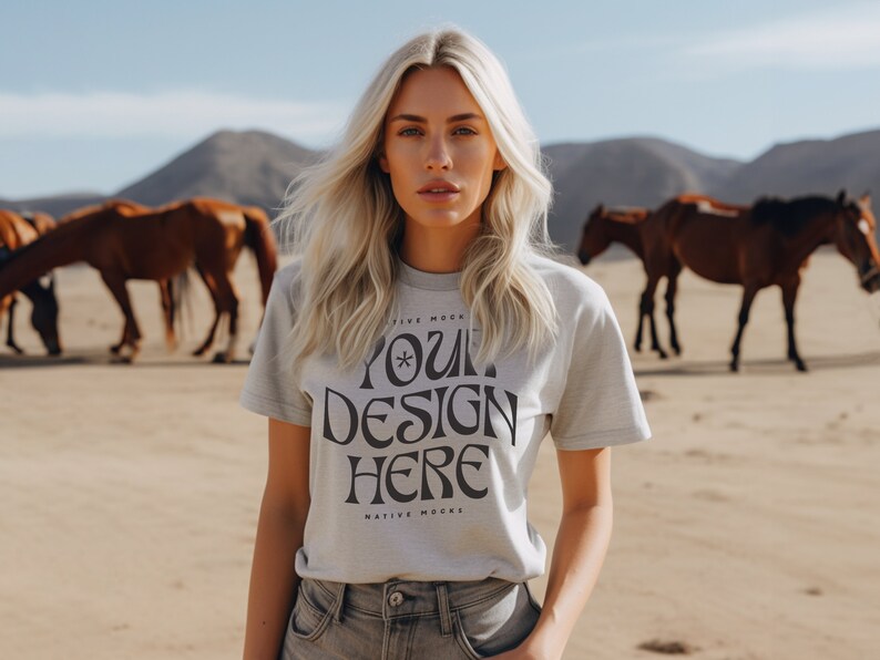May include: A woman wearing a light gray t-shirt with the text "YOUR DESIGN HERE" printed on it. The shirt is a mockup for a design project. The woman is standing in a desert landscape with horses in the background.